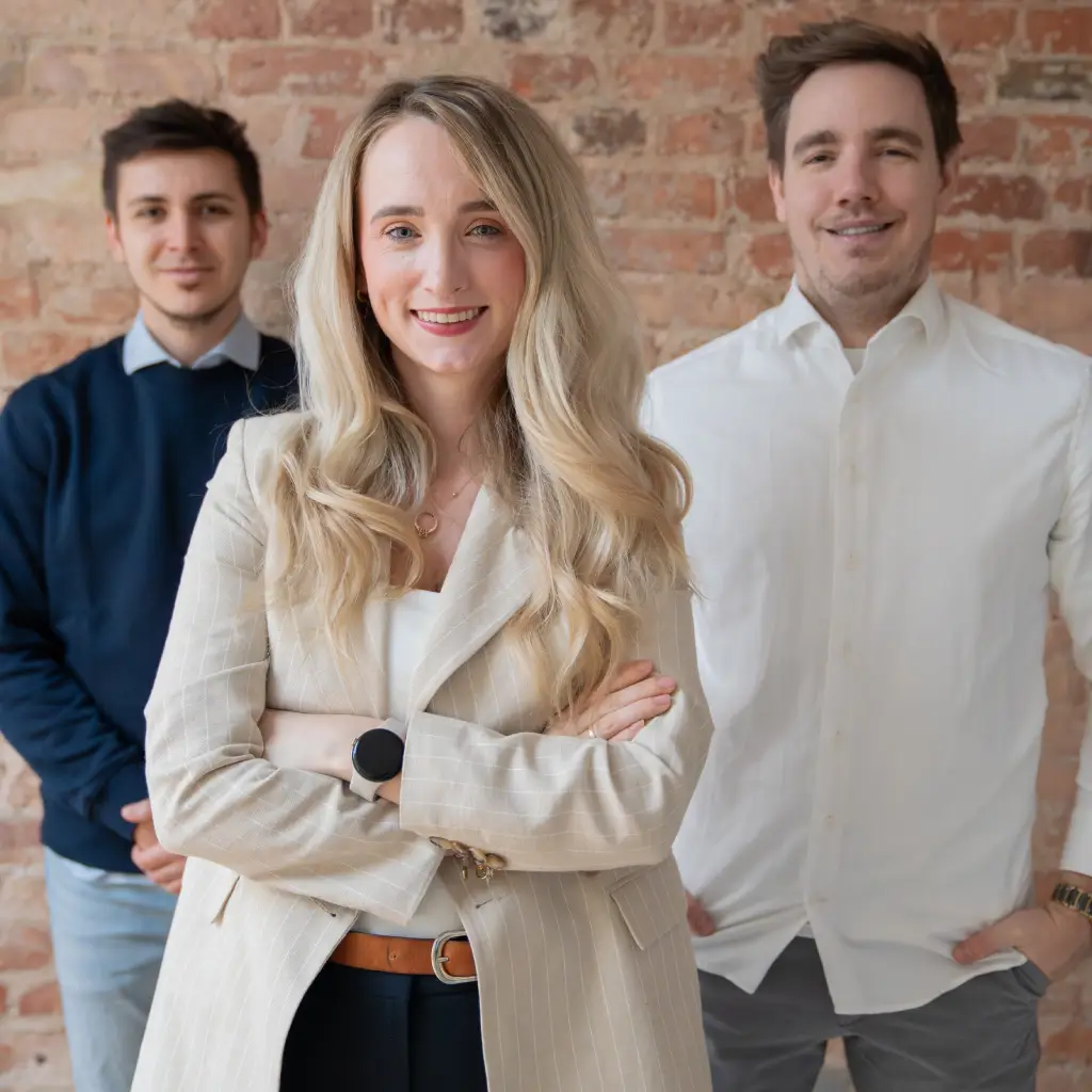 Drei junge Erwachsene vor einer Backsteinwand, eine Frau im Vordergrund, zwei Männer im Hintergrund.