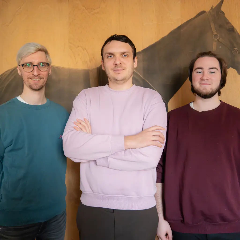 Drei junge Männer stehen vor einer Wand mit Pferdebild, lächelnd, lässig gekleidet.